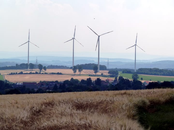 Vier Windkraftanlagen in der Landschaft mit Feldern, auf einer Anhöhe. Es ist etwas diesig.